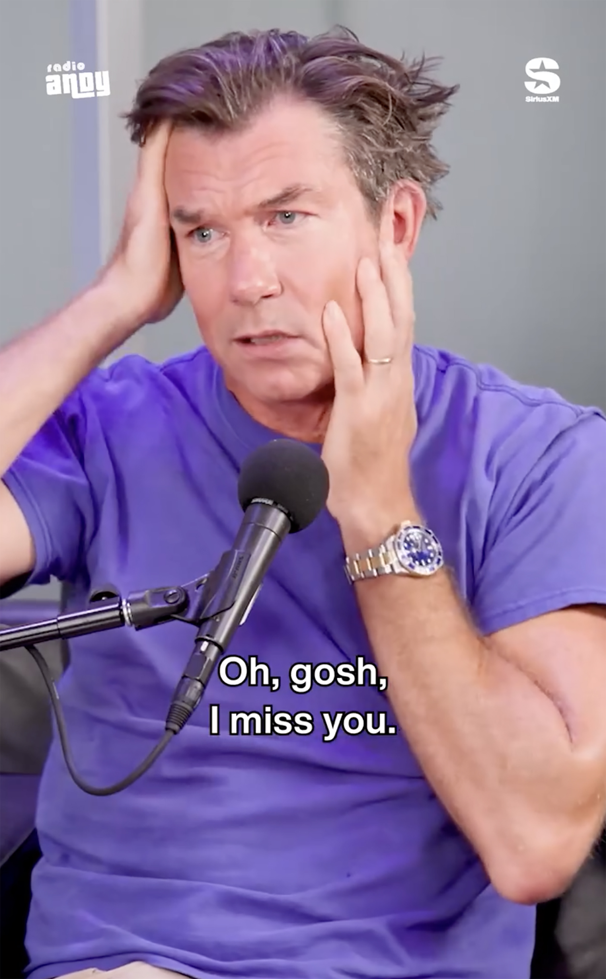 Jerry O'Connell with purple-streaked hair, holding his head with hands, and a microphone in front of him.