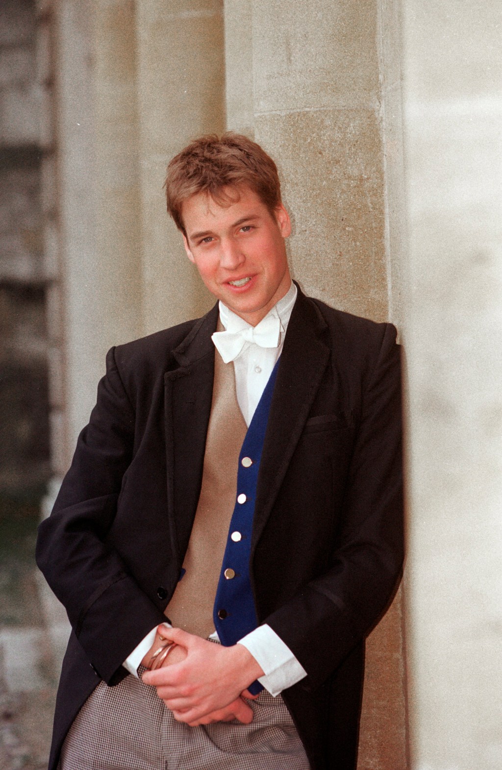 Prince William in a formal school uniform, including a tailcoat, bow tie, and a blue waistcoat, leaning against a stone wall.