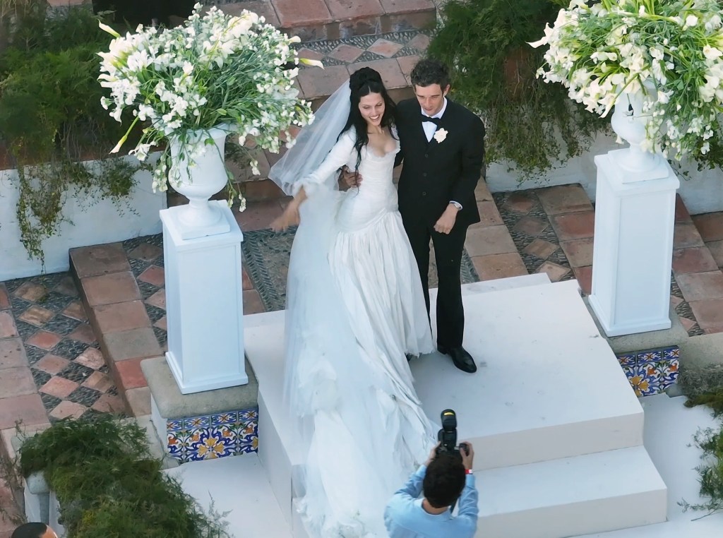Matty Healy and Gabbriette Bechtel walk down wedding aisle, a photographer in the foreground.