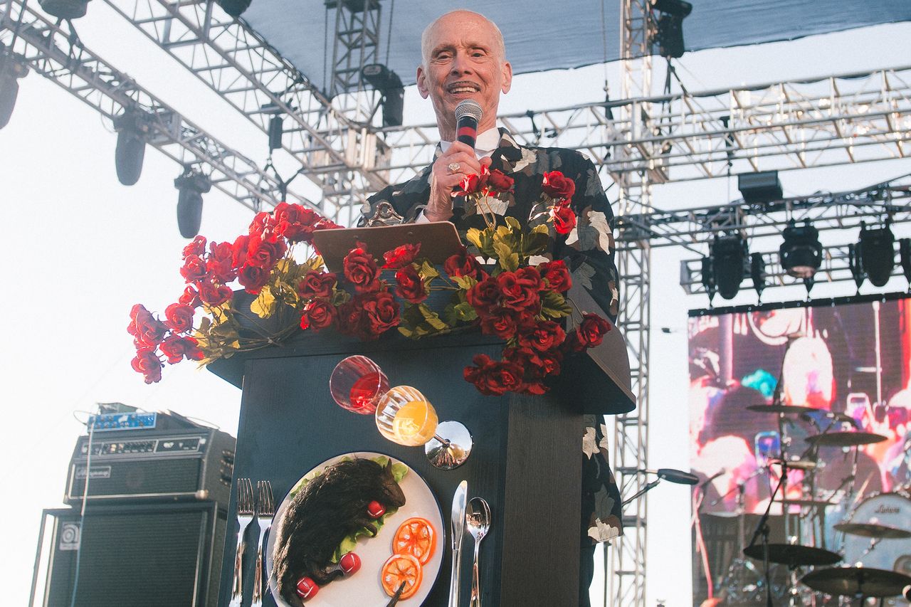 John Waters speaking at a decorated podium on an outdoor stage