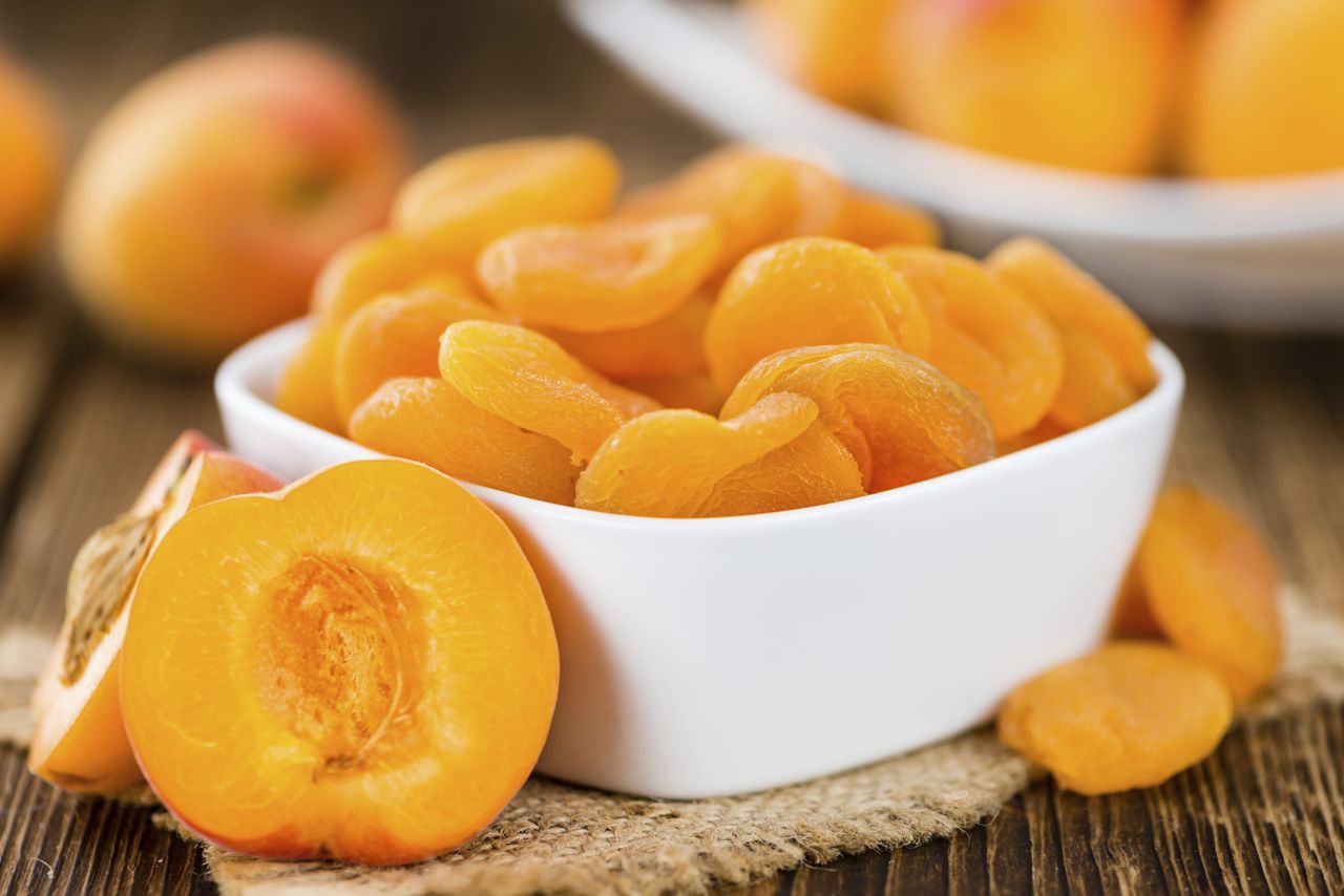 A bowl of dried apricots and some fresh apricot halves on a wooden surface