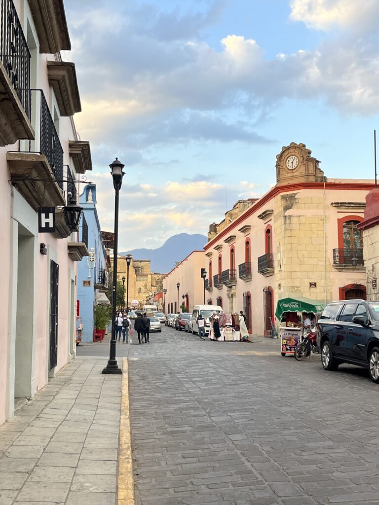 A quiet cobblestone street in Oaxaca is lined with pastel colonial buildings, wrought-iron balconies, and vintage streetlamps, with mountains visible in the distance beneath a sky of soft blue and golden clouds. A historic clock-topped building and small street vendors add to the city's timeless atmosphere.