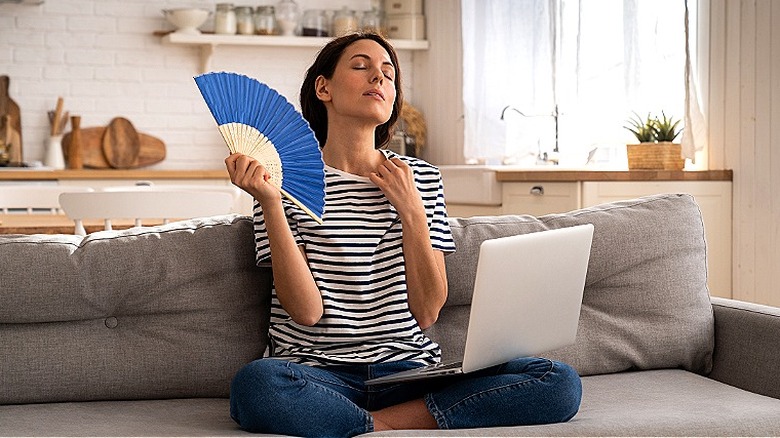 A person in a striped shirt sits on a couch with a laptop while using a blue hand fan to cool down