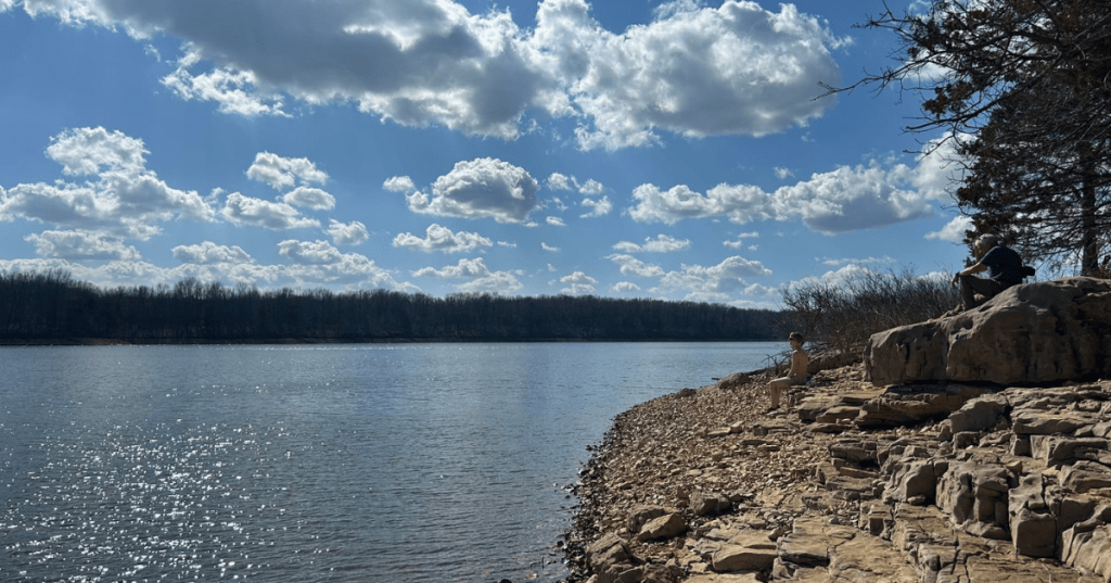 People relaxing on the rocky shore of Fellows Lake in Missouri under a bright, cloudy sky.
