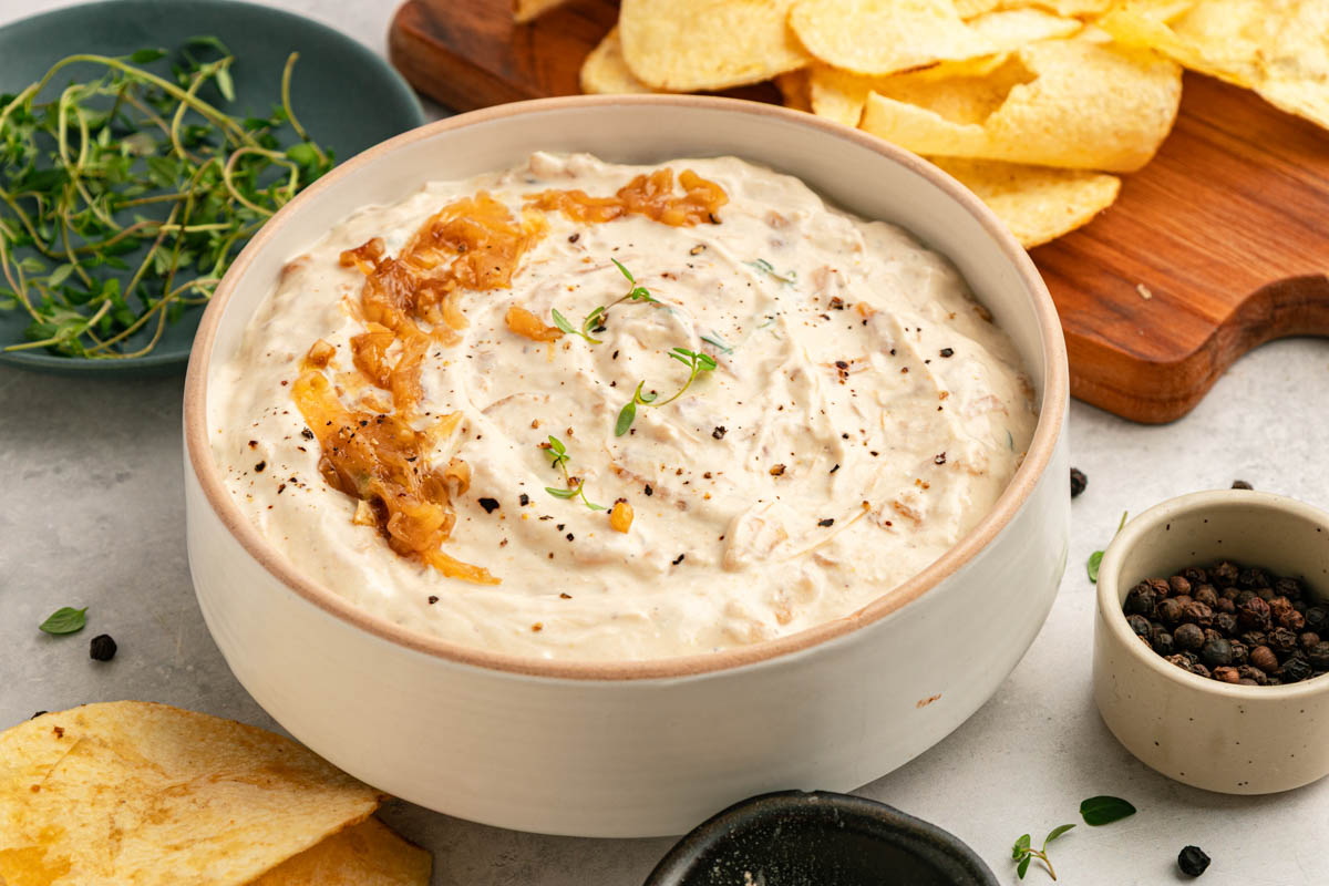 Bowl of creamy dip garnished with caramelized onions and herbs, surrounded by potato chips, peppercorns, and fresh greens.