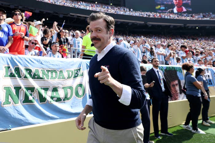 Person at a sports event pointing, wearing a sweater over a shirt, surrounded by a cheering crowd
