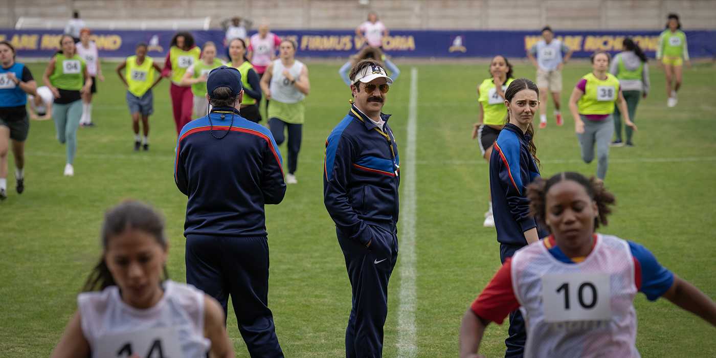 Ted looking back with other coaches while the girls practice on the field in Ted Lasso.