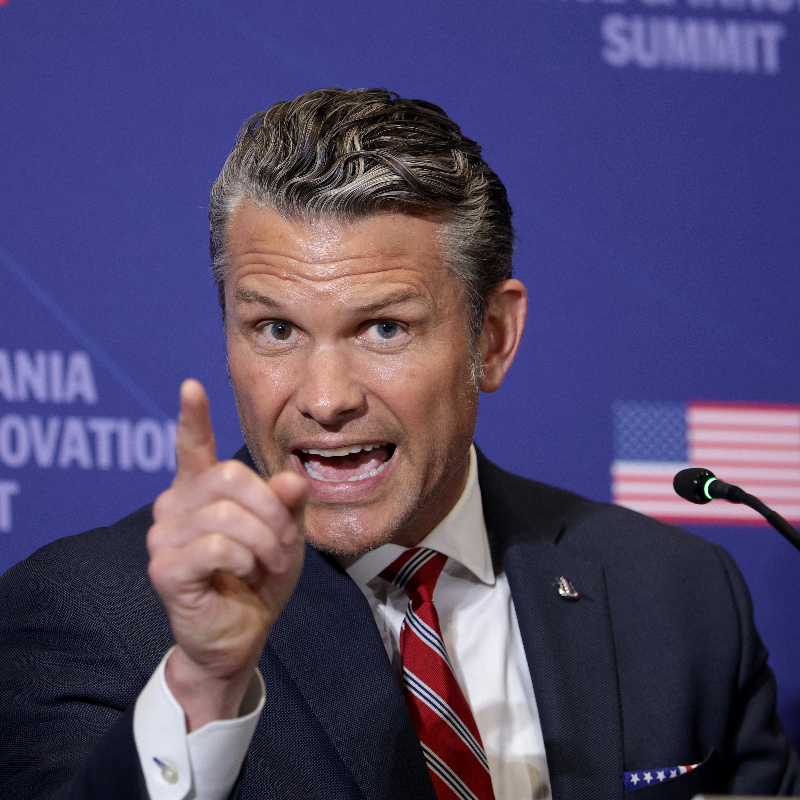 A man in a suit gestures emphatically while speaking at the Pennsylvania Defense & Innovation Summit, with an American flag in the background