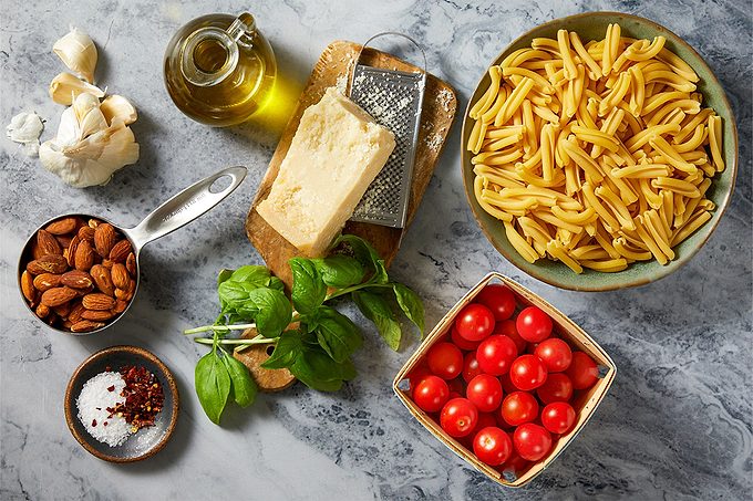 Overhead horizontal image of various fresh ingredients laid out on a marble surface. The arrangement includes garlic, a cruet of olive oil, a block of Parmesan cheese with a grater, and a bowl of pasta. Also visible are a measuring cup full of almonds, fresh basil, cherry tomatoes, and a dish with salt and red pepper flakes.