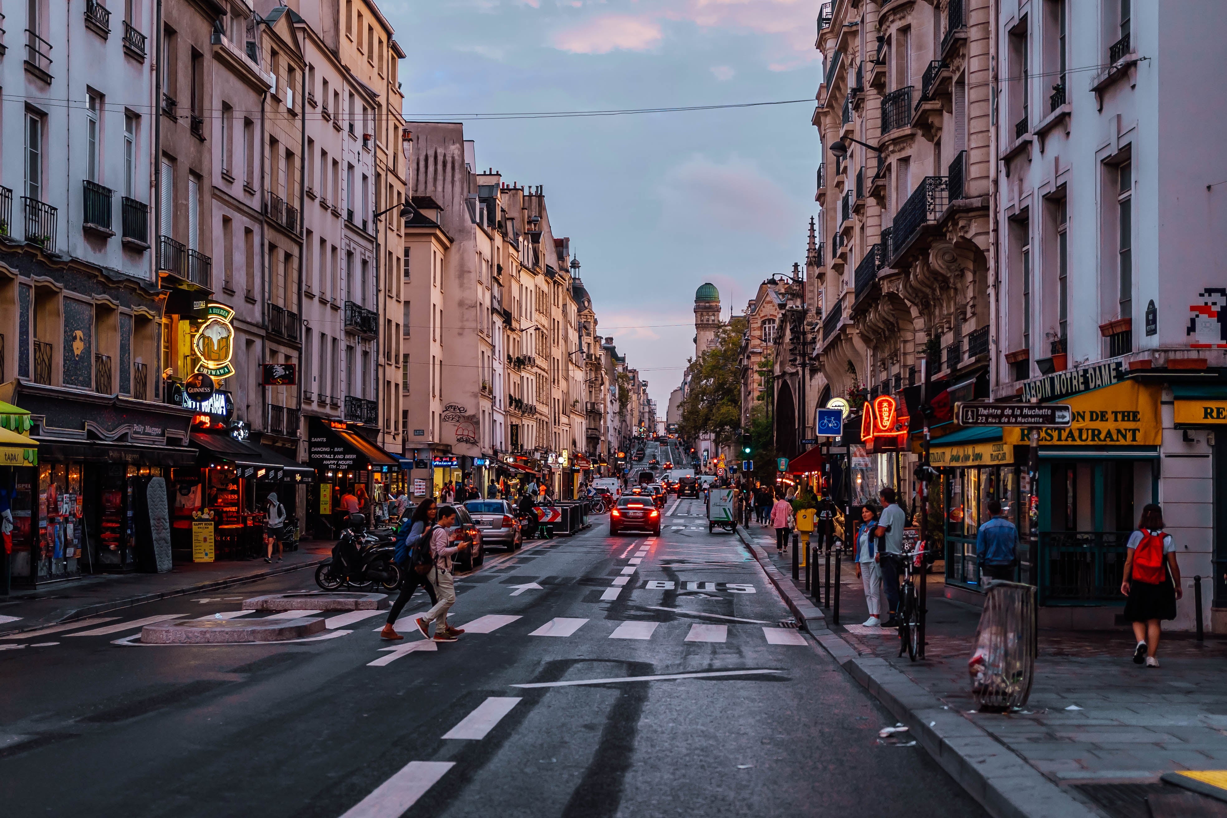 A bustling Paris street with people walking, cars, bicycles, and neon signs. Historic buildings line the street, capturing a lively urban scene