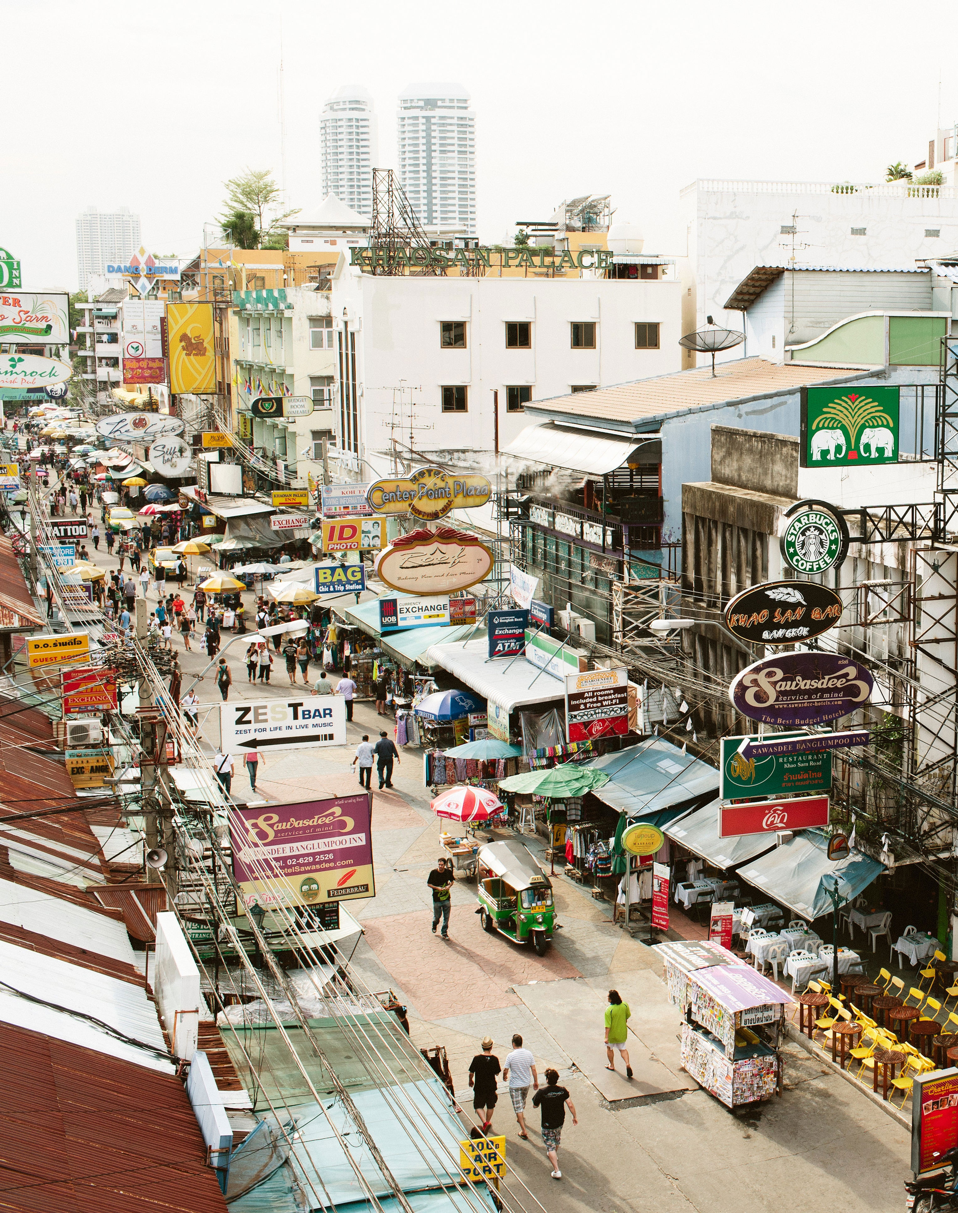 Busy street market with people walking, various shop signs, and vendors selling goods, capturing vibrant local life likely in a Southeast Asian city