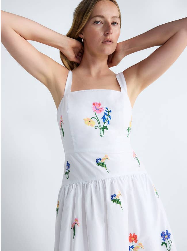Person in a sleeveless, floral-embroidered sundress poses with arms slightly raised, showcasing the dress's fit and detailing