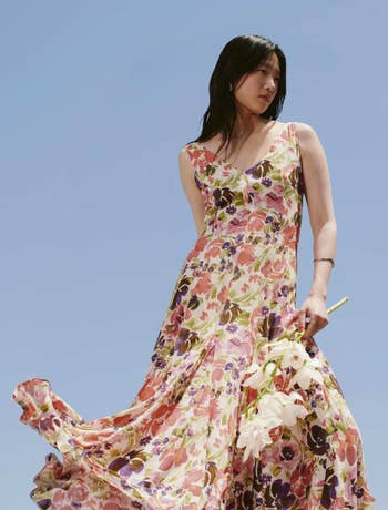 Person in a flowing floral dress stands confidently against a clear sky, holding white flowers, showcasing a spring fashion style