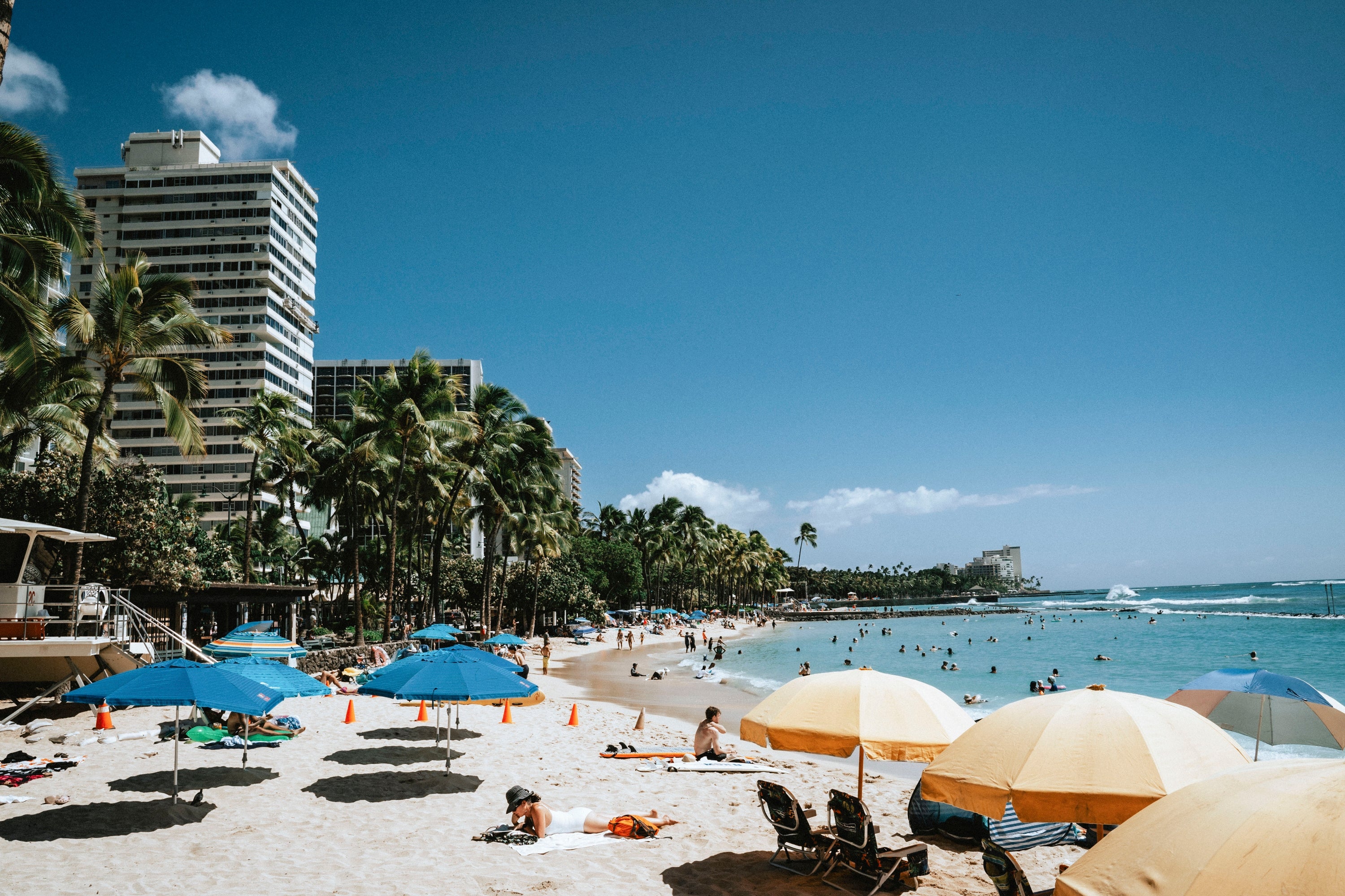 Beachgoers enjoy a sunny day at a bustling oceanfront, with palm trees and high-rise buildings in the background