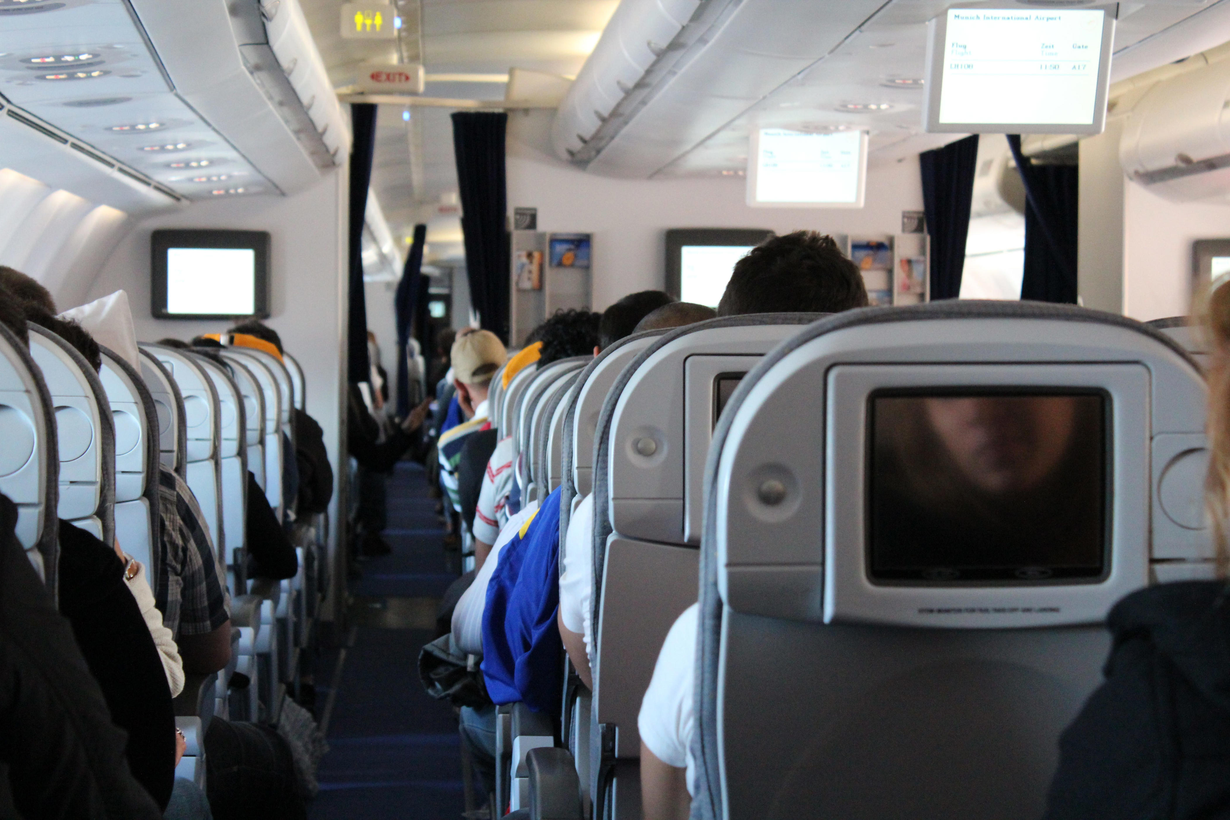 Passengers sit in an airplane cabin with rows of seatback screens and overhead monitors visible.