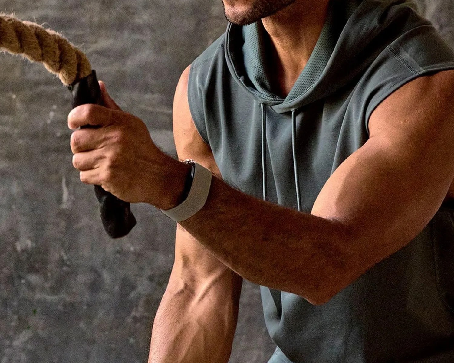 Close-up of a muscular arm in a sleeveless gray hoodie holding a thick rope with a black grip.