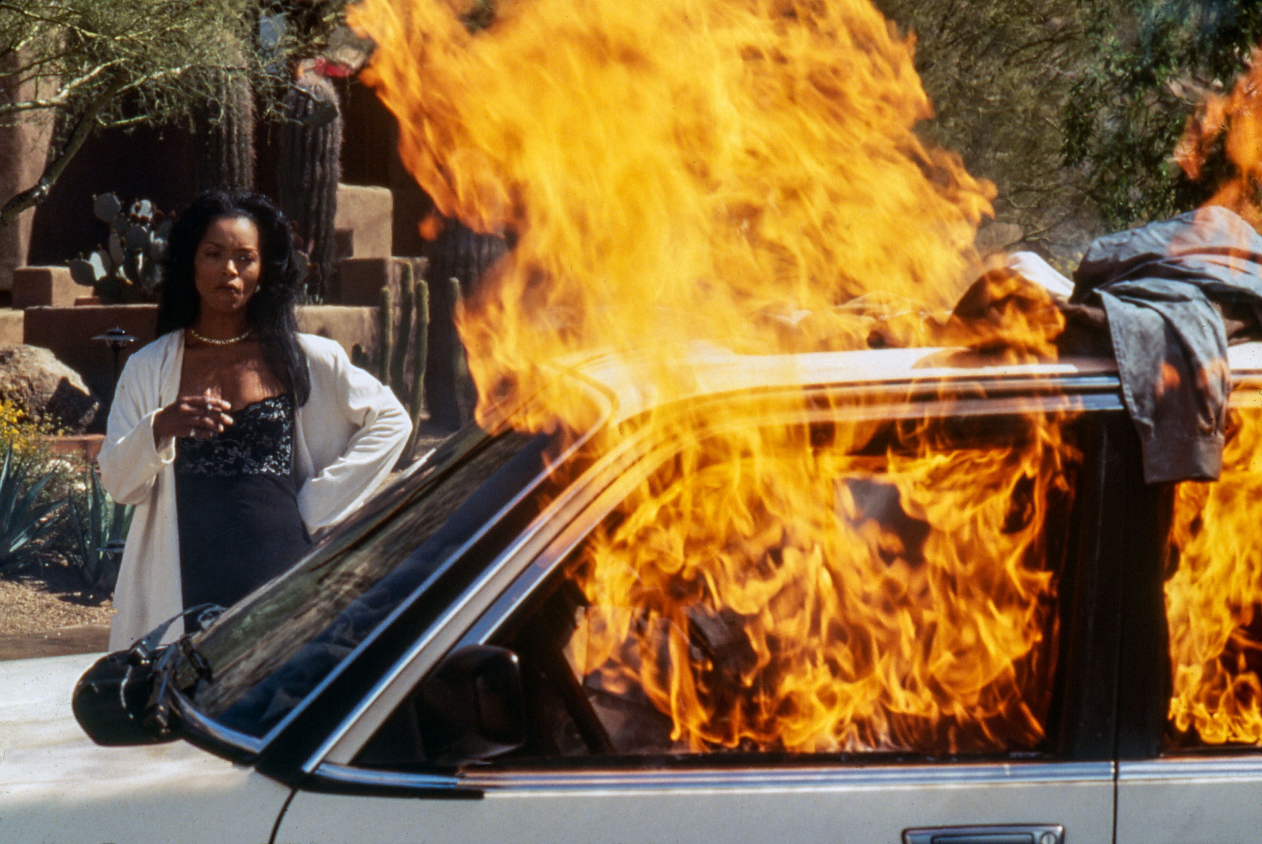 A woman in fashionable attire stands confidently beside a burning car outdoors