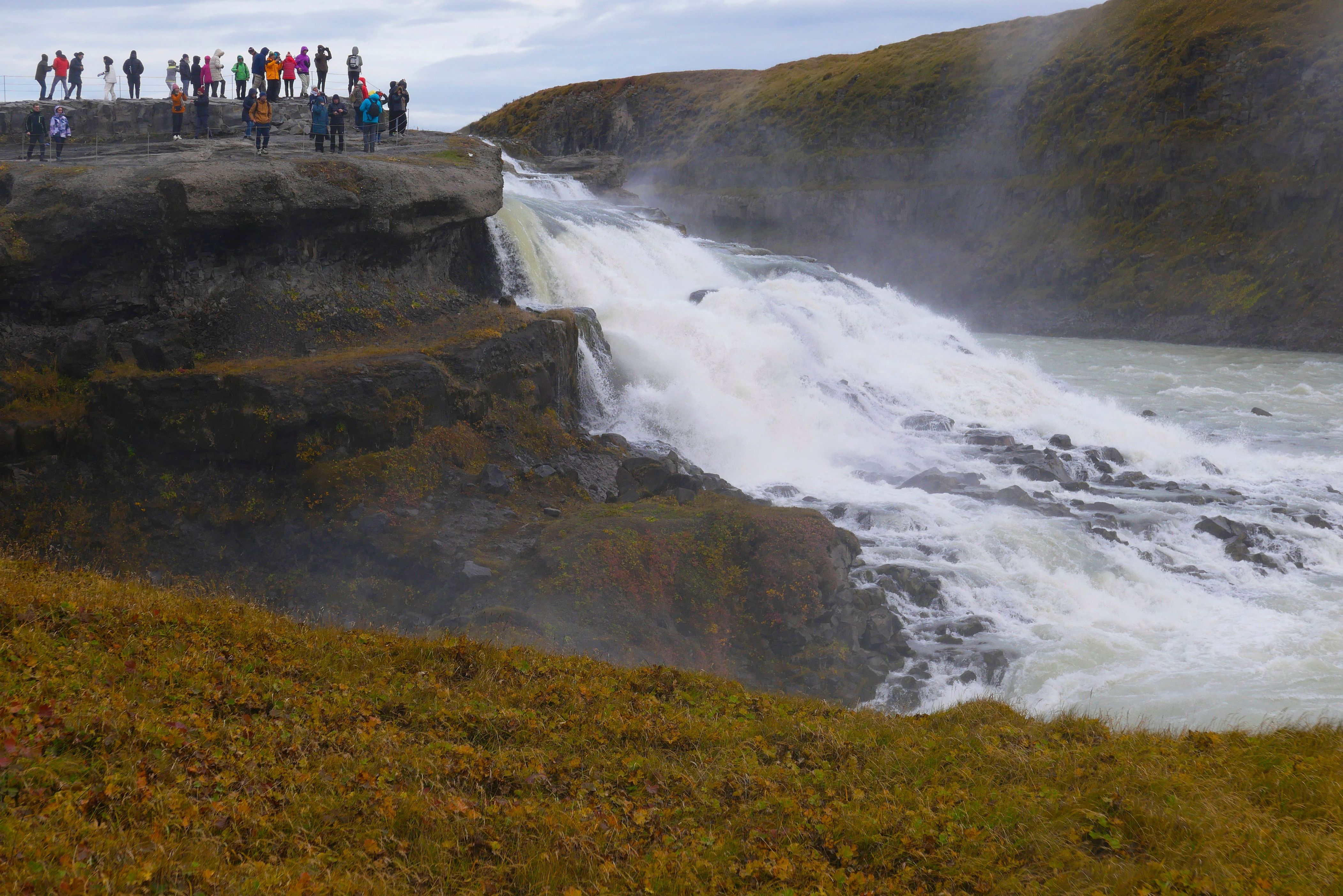 Tourists stand on a cliff overlooking a large waterfall cascading into a rocky gorge, surrounded by grassy hills