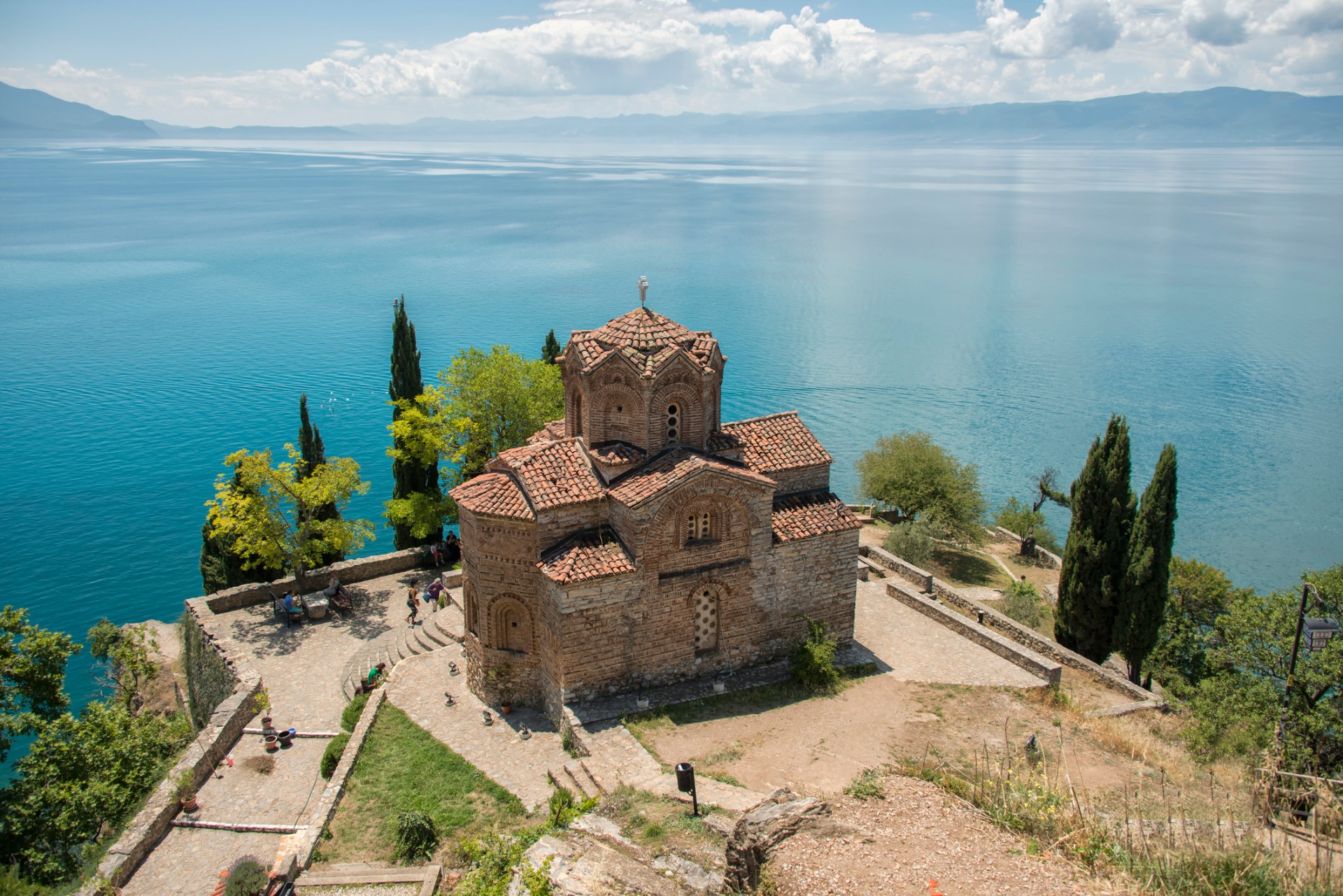 The Orthodox church of Saint John the Theologian overlooking Lake Ohrid. The church has a brown, terracotta roof and the lake is a dazzling shade of blue. Trees line the cliff above it.