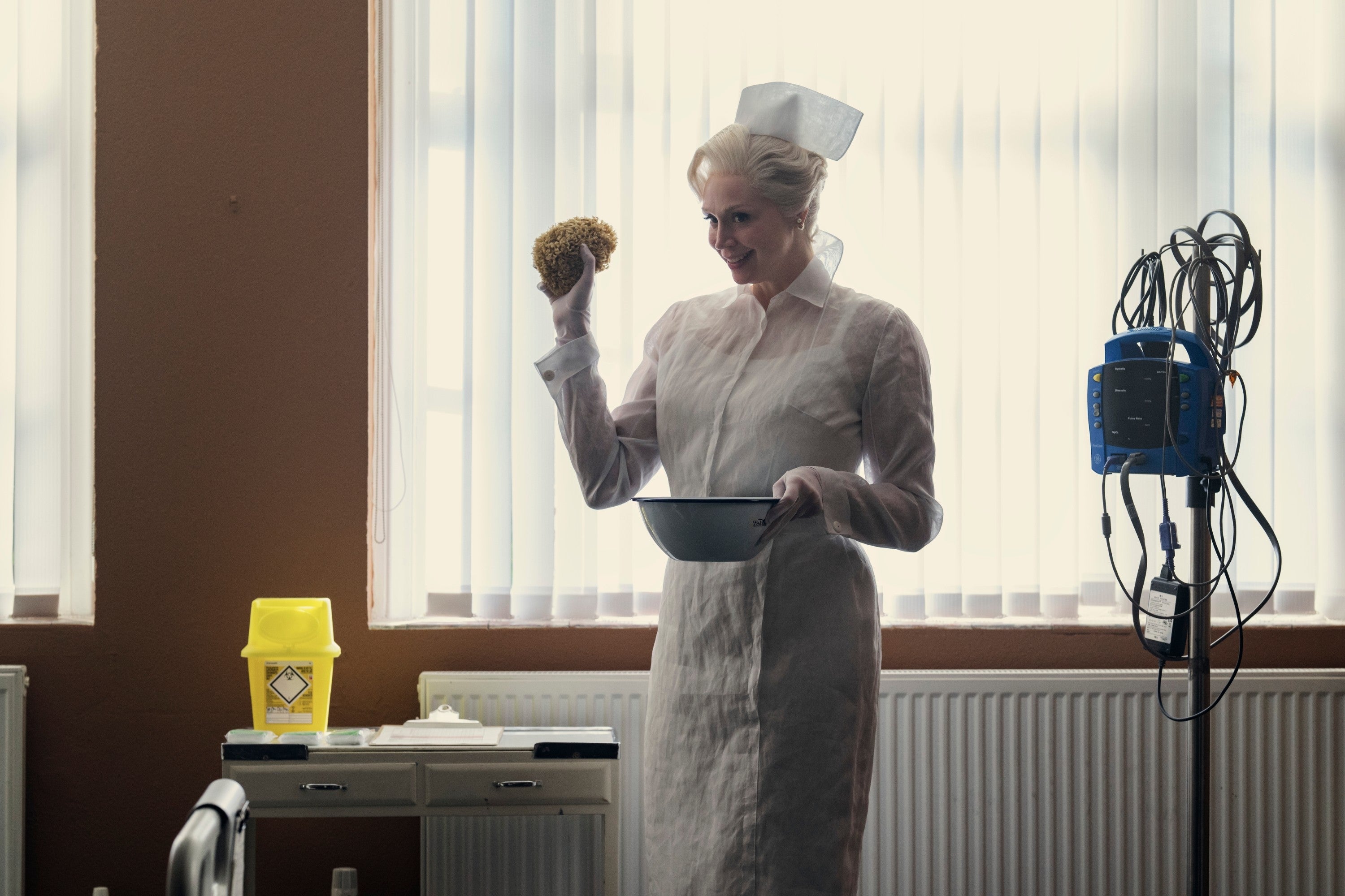 Person in vintage nurse attire holding a bowl and a sponge in a hospital setting. Medical equipment is visible nearby