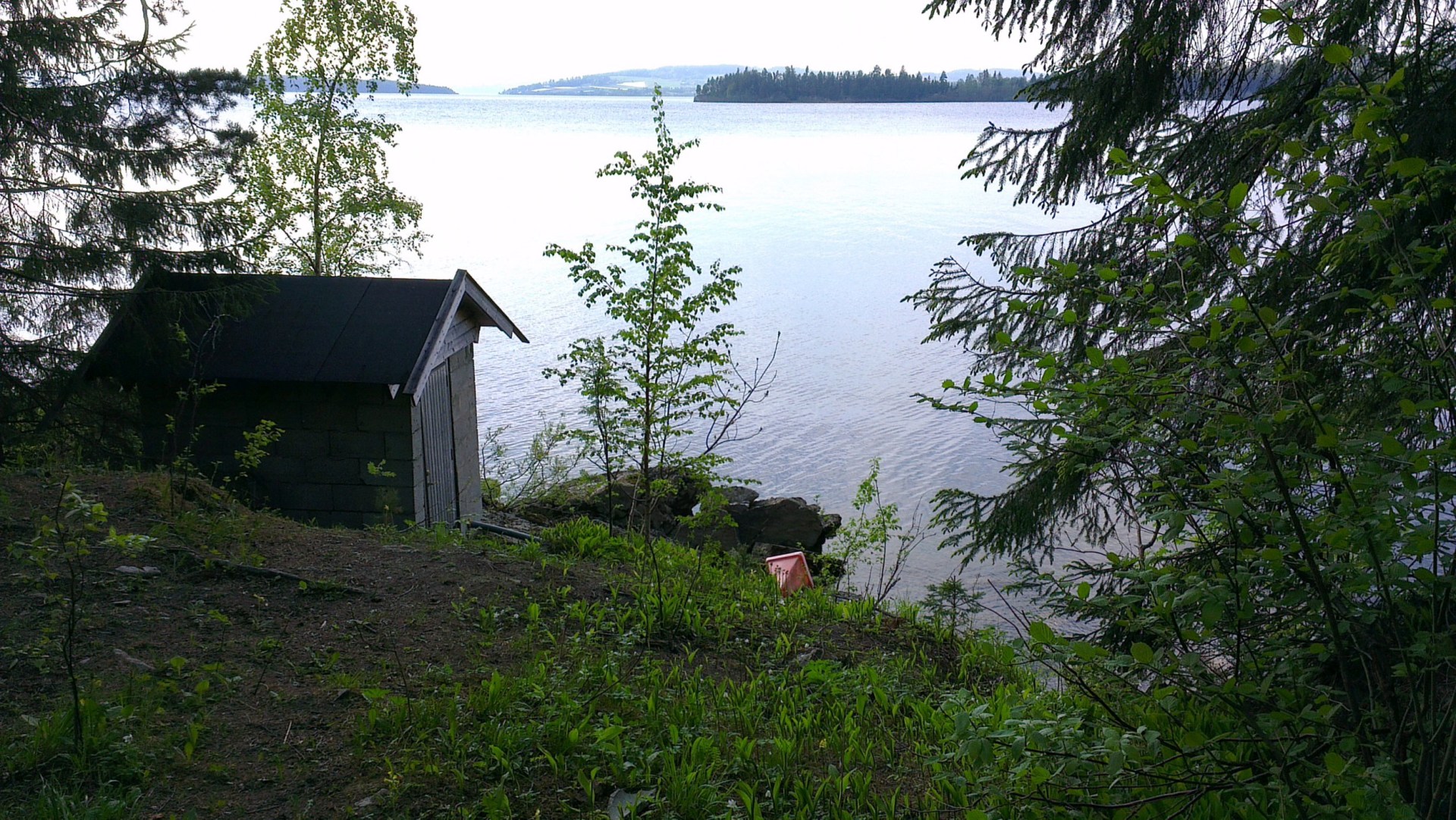A small shed by a large body of water. Thhere are trees and there is grass.