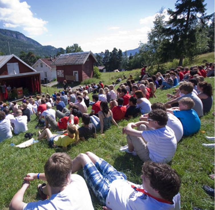 UTOYA YOUTH CAMP. Many children sit on grass by traditional Norwegian buildings.