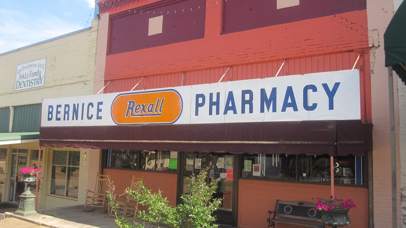 A white and orange sign for 'BERNICE Rexall PHARMACY' hangs above a storefront with a dark brown awning. The building is made of orange brick with a darker red upper section. Outside, several wooden rocking chairs, a dark metal bench, and a planter with vibrant pink flowers are visible. To the left, a cream-colored building has a sign for 'Kurt Touchstone DDS ArkLa Family DENTISTRY', all under a clear blue sky.