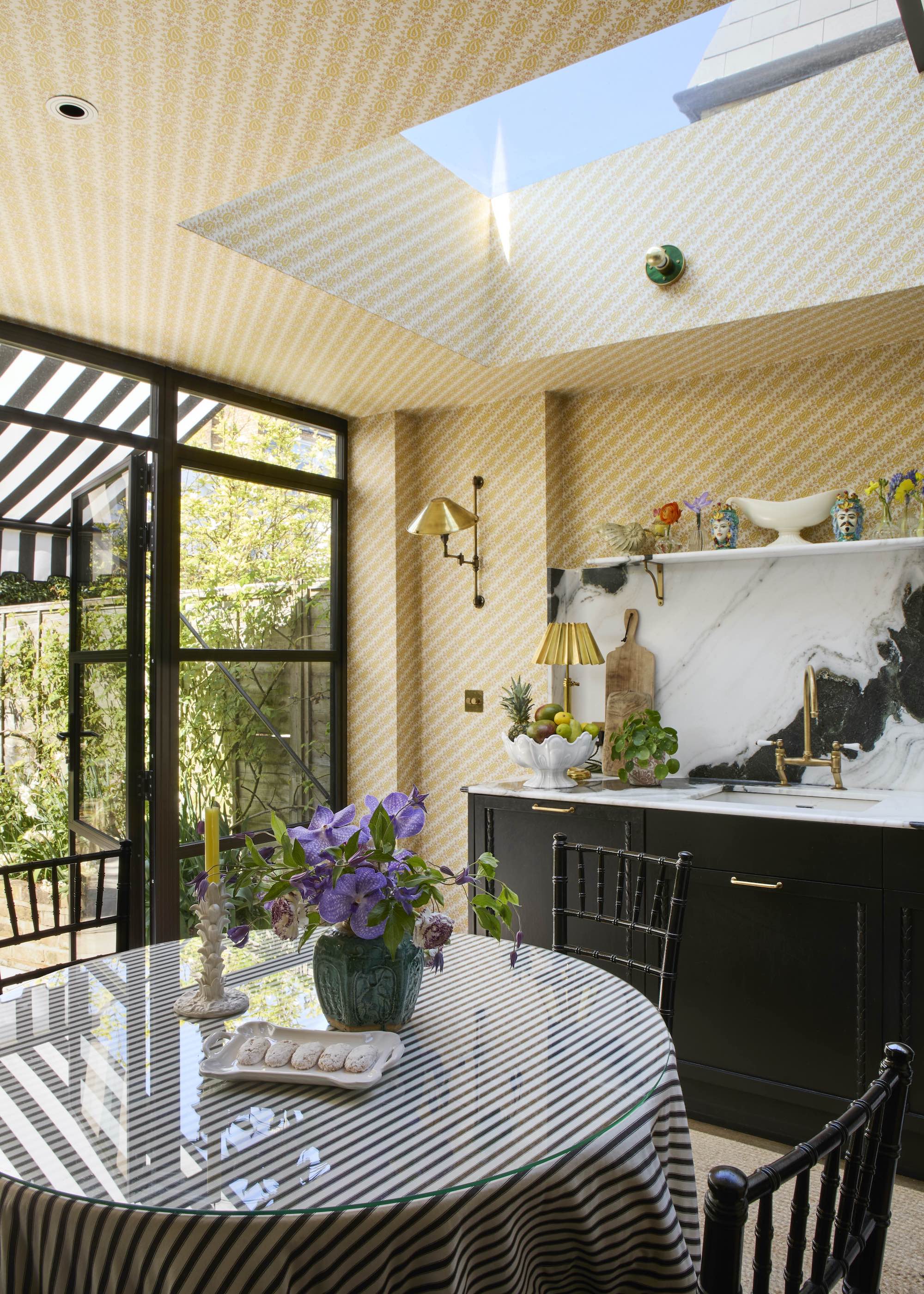 black kitchen with striped yellow wallpaper on the walls and ceiling and skylight, with white marble backsplash and shelf, with round glass table in the foreground with black striped tablecloth