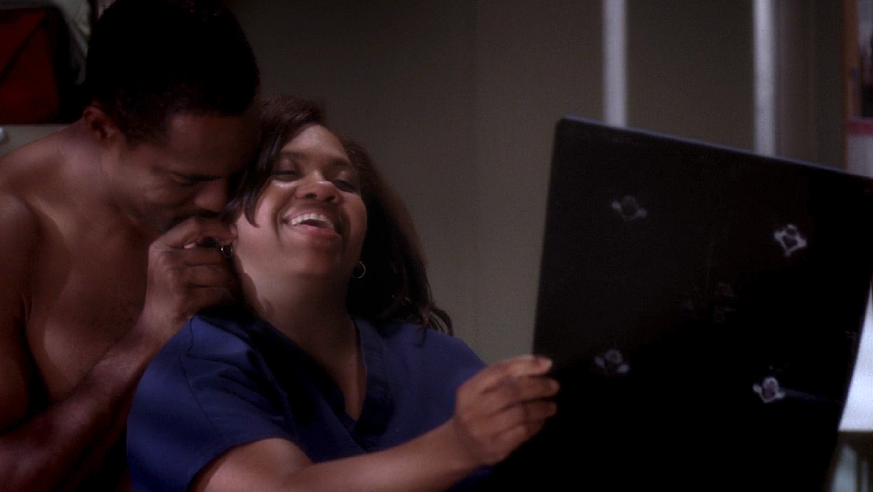 A person in scrubs holds a tablet, laughing joyfully with another person embracing them warmly from behind