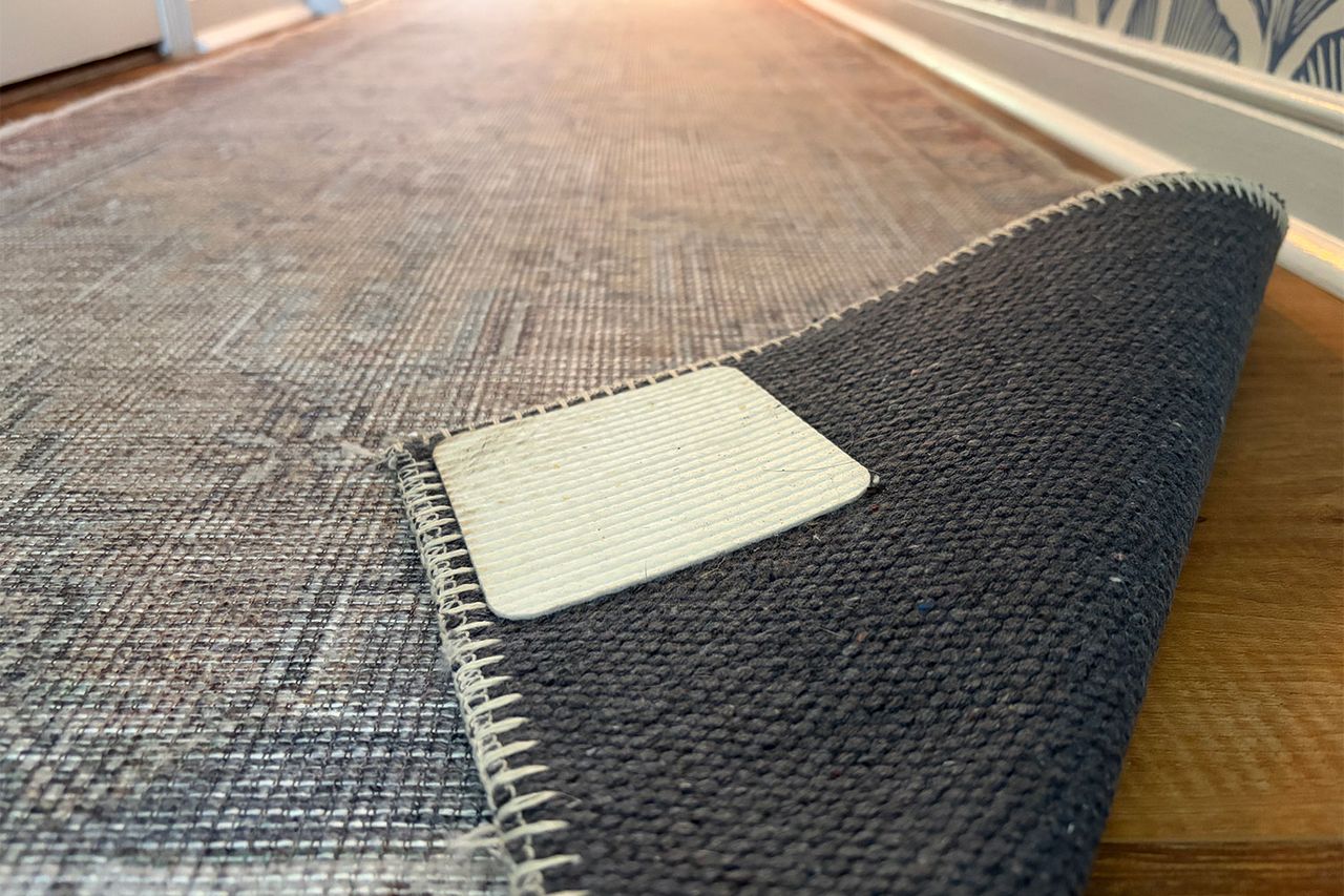 A rug with a corner lifted to show a rug pad and gripper underneath on a wooden floor hall in background