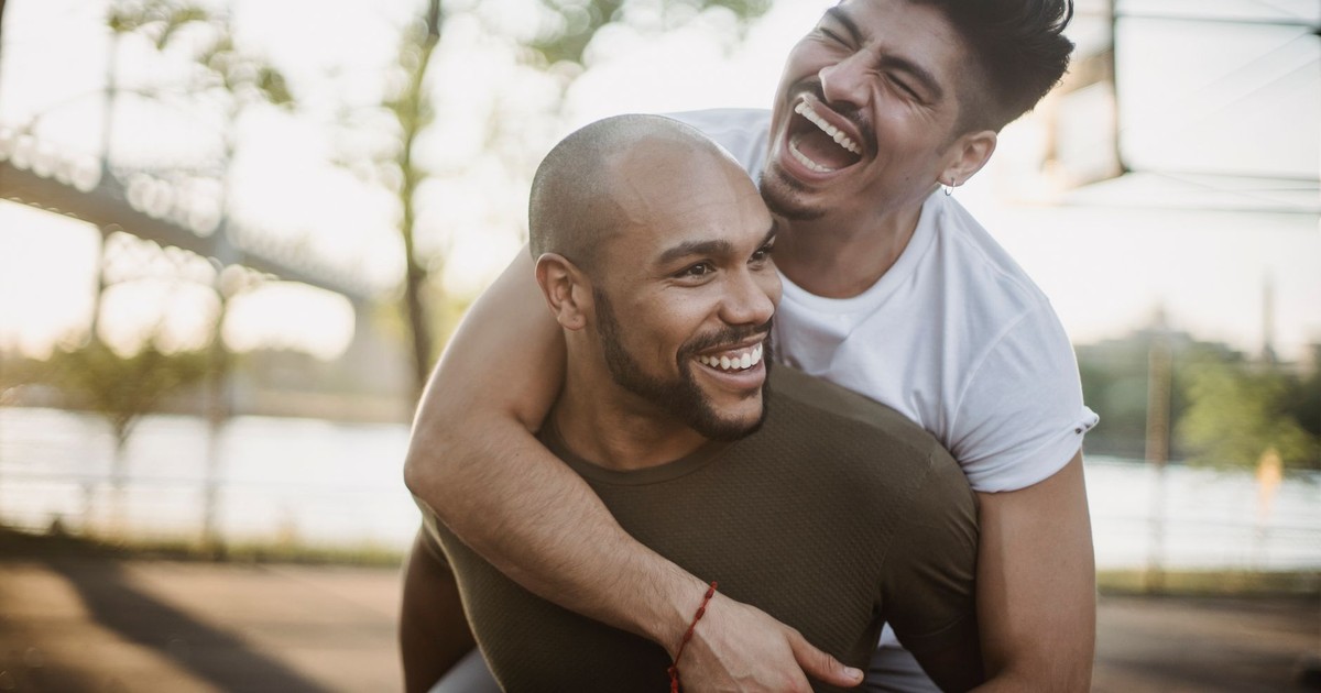 Happy interracial gay couple enjoying a playful piggyback ride in a sunny park setting.