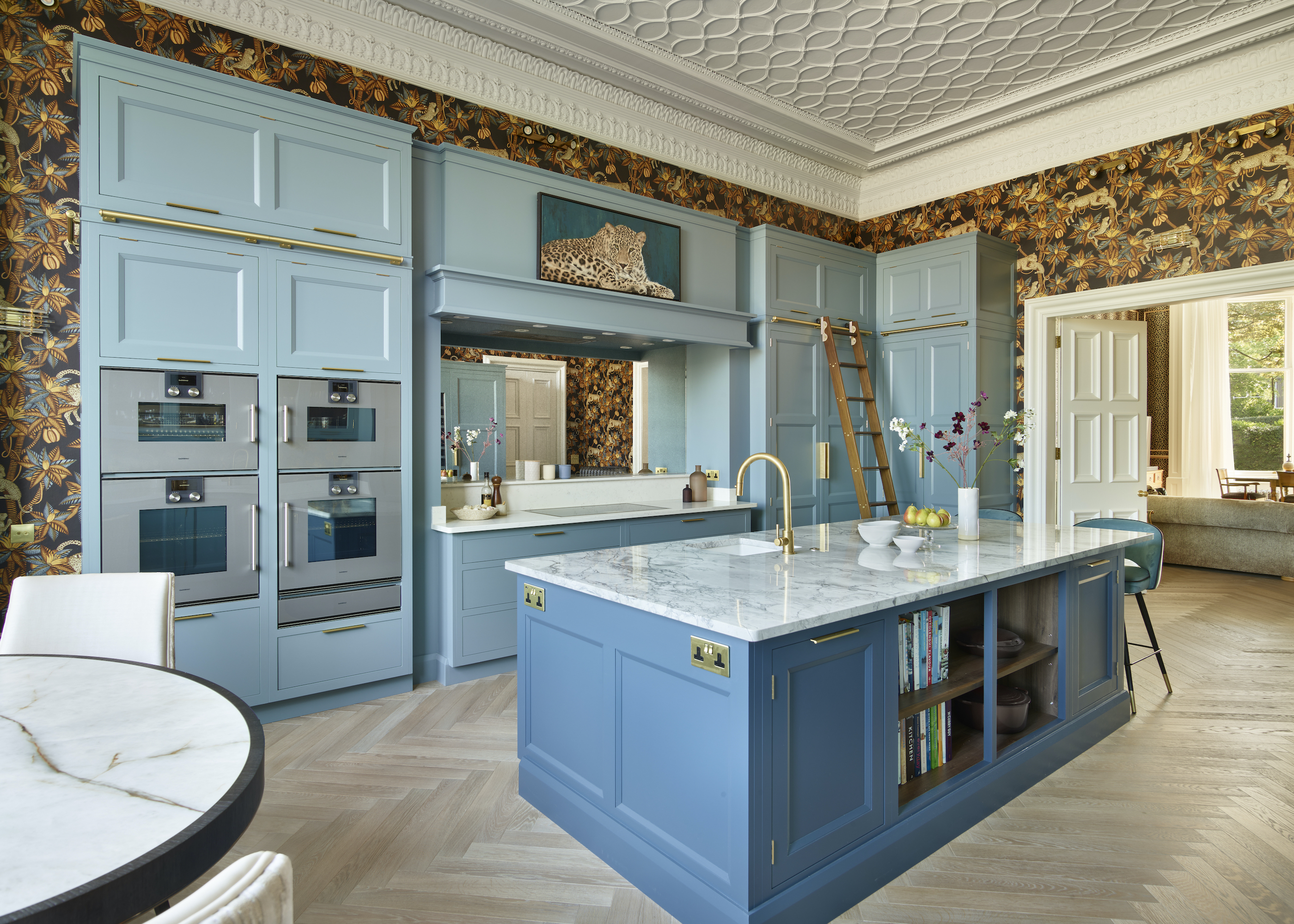 blue kitchen with exotic wallpaper, pressed metal ceiling, double appliances, blue kitchen island with stone countertops, rolling ladder, brass taps, round table with white chairs