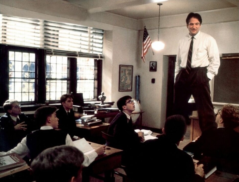 A man standing in front of a classroom speaks to students seated at desks near large windows.