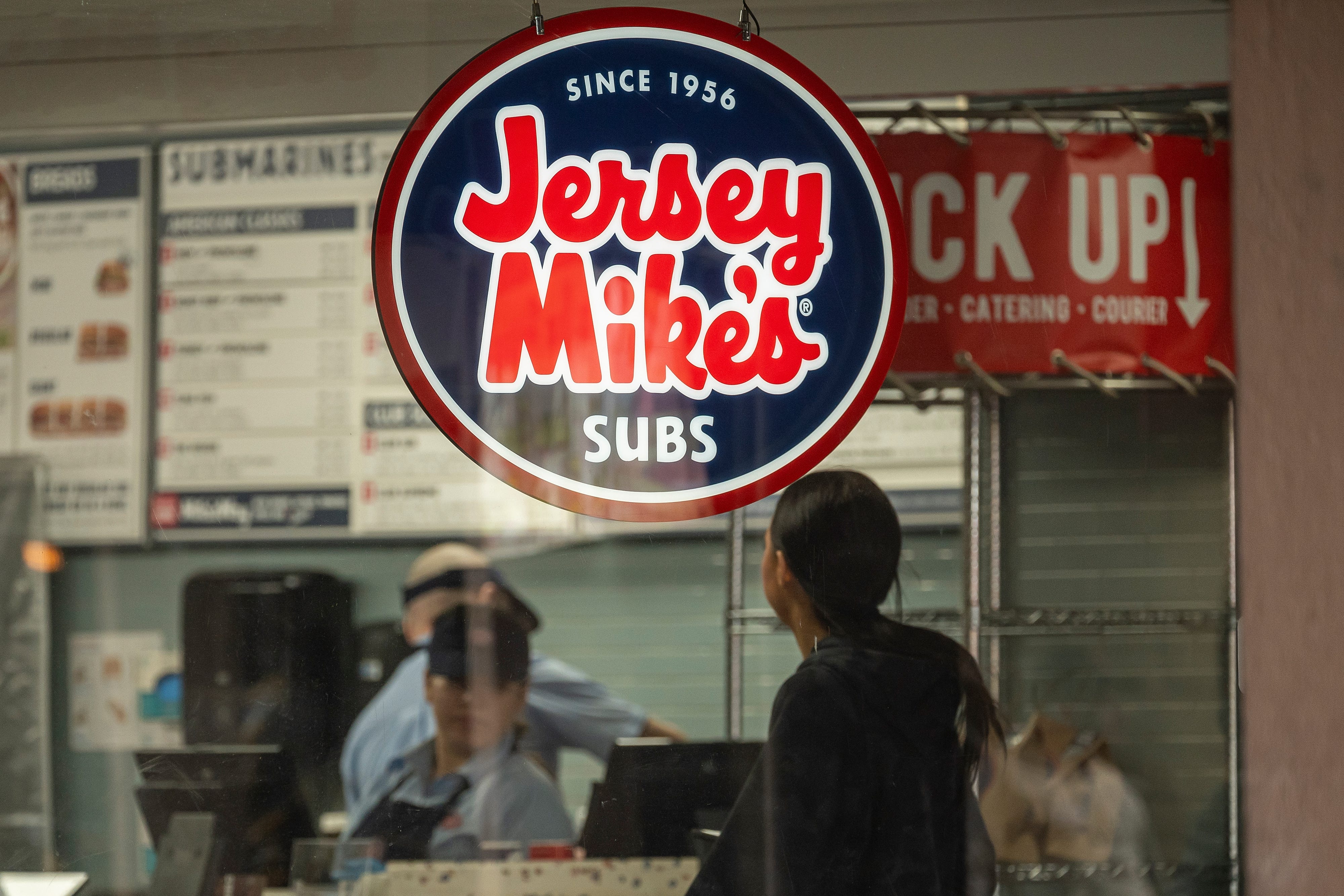 A red, white, and blue lighted Jersey Mike's sign hangs in the window of a restaurant as a customer stands at a counter and store employees wear hats and aprons behind it.