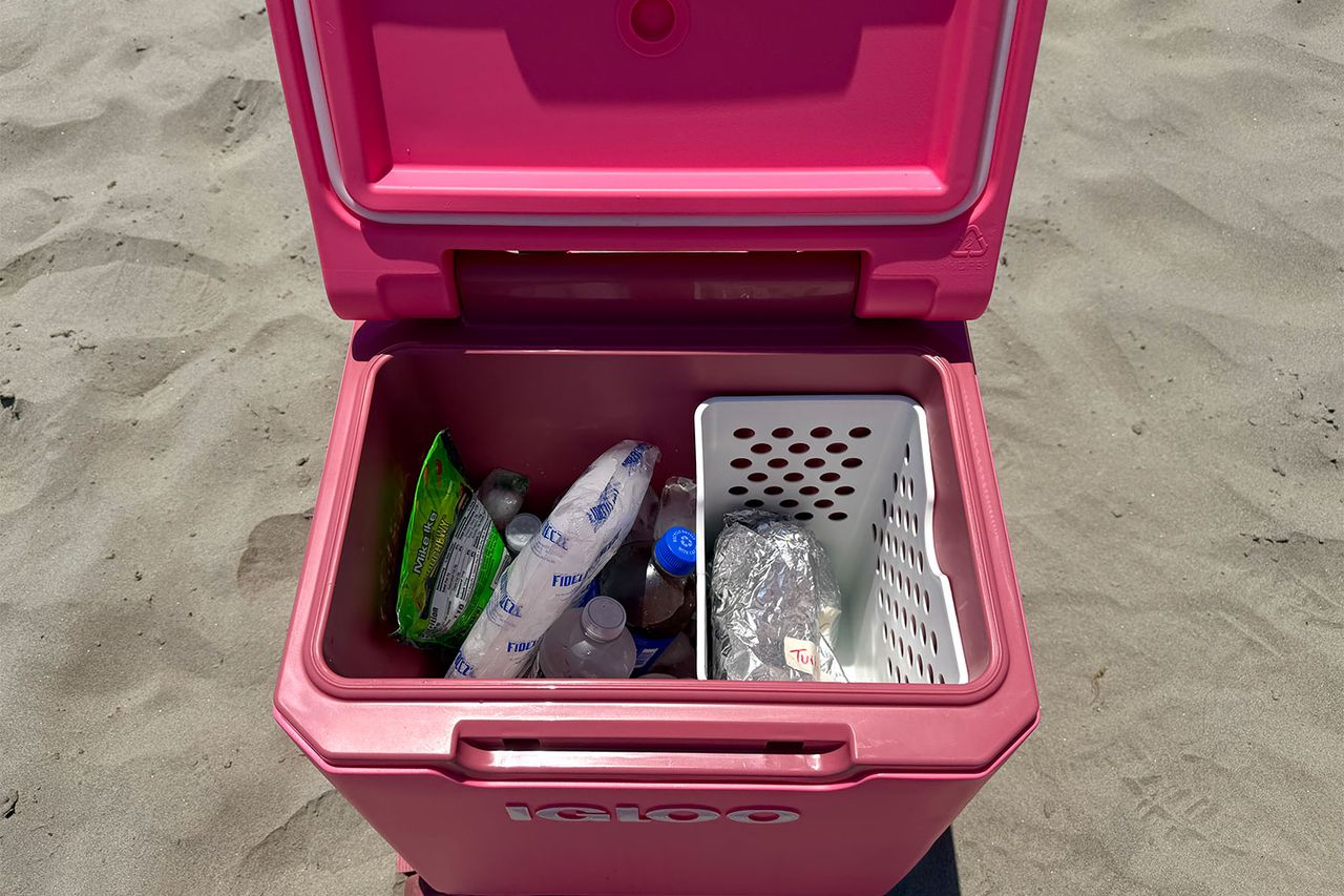 An Igloo brand cooler with contents including ice packs bottles and containers placed on a sandy surface