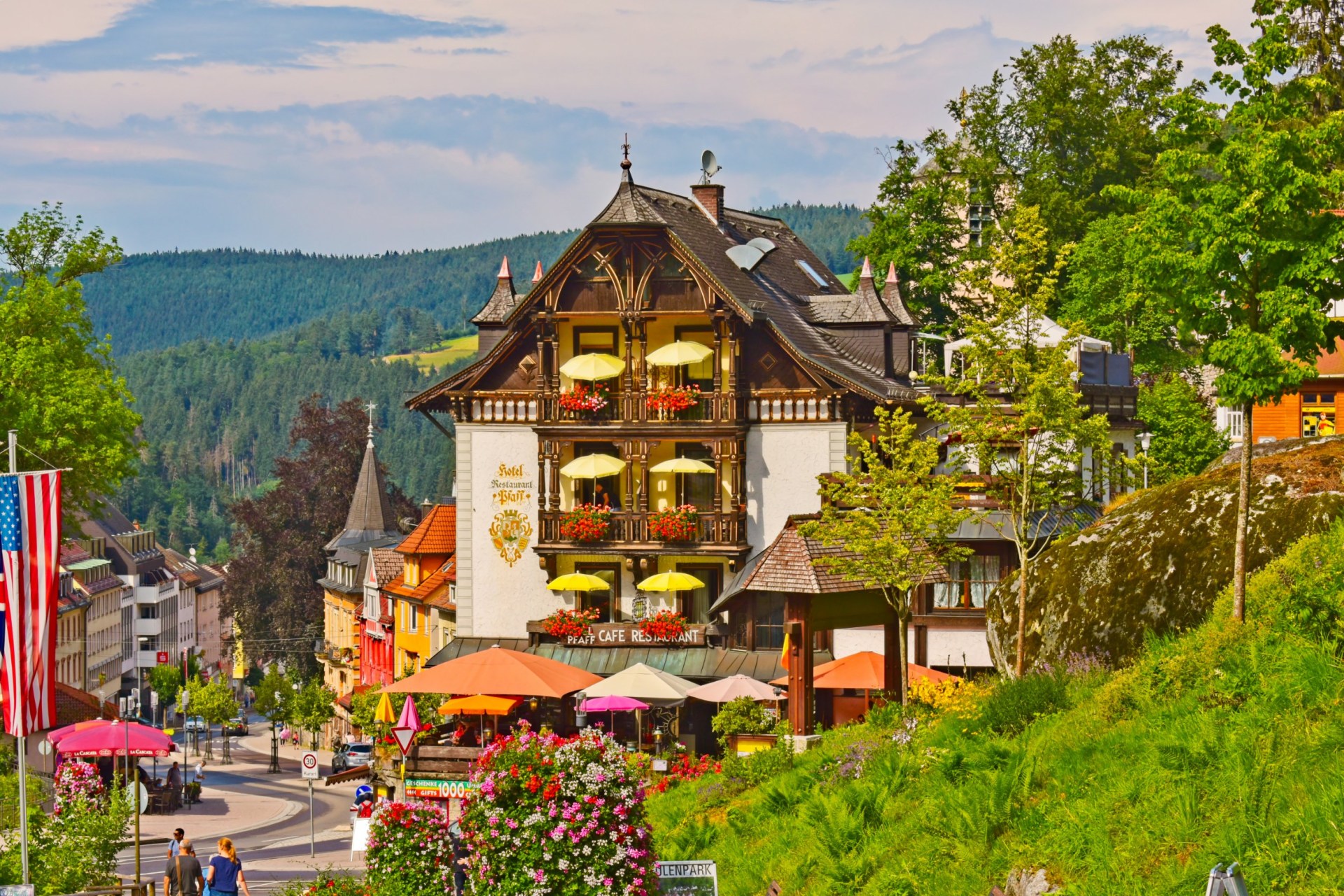 A hotel in Triberg, Germany, surrounded by greenery. There are umbrellas on the hotel's balconies and seating areas below.