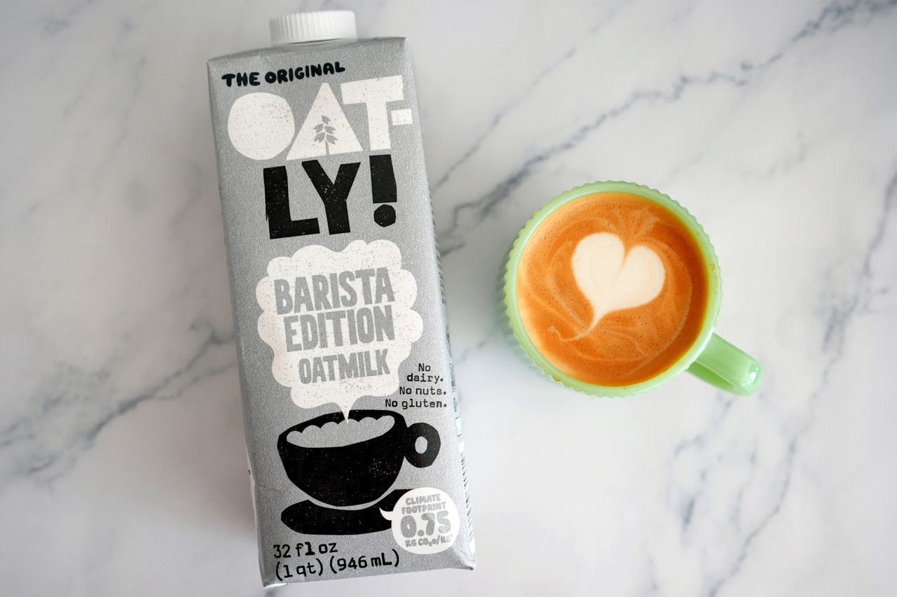A carton of Oatly Barista Edition oatmilk beside a cup of latte with heartshaped foam art