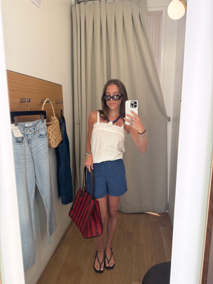 Person in a dressing room wearing summer attire and holding a phone standing near hanging jeans and bags