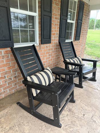 Two wooden rocking chairs with striped cushions on a porch, positioned in front of a brick wall with windows, creating a cozy outdoor setting