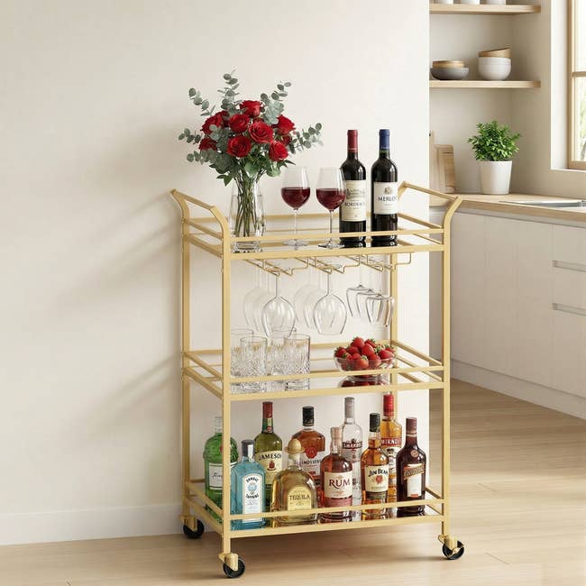 Gold bar cart in a kitchen, stocked with wine, spirits, glasses, and a vase of red roses
