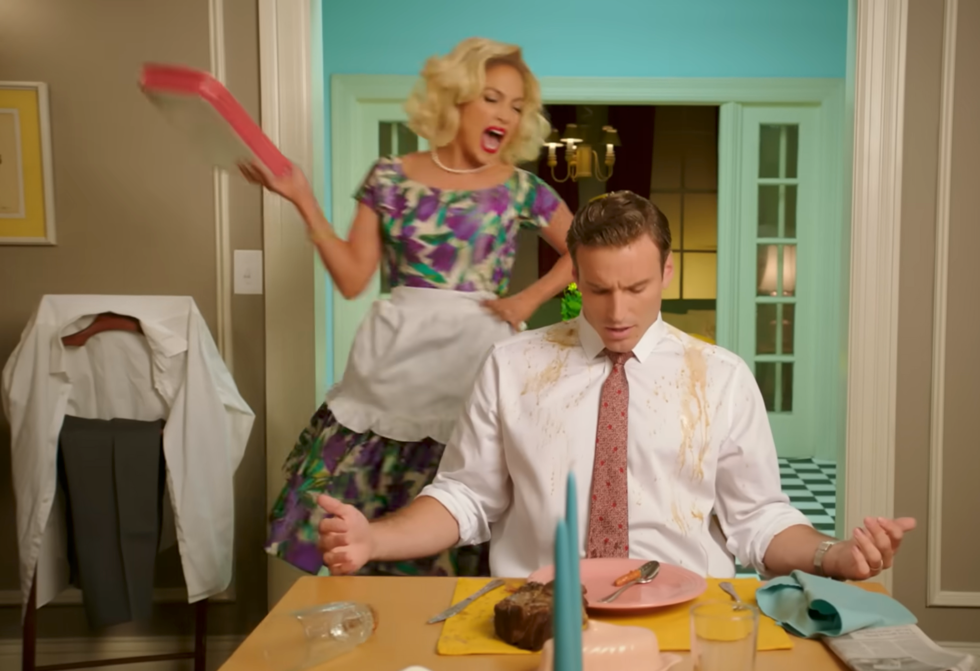 A person in retro-style clothing, with an apron, appears to angrily yell while holding a spatula. Another seated person looks annoyed, with food on their clothes