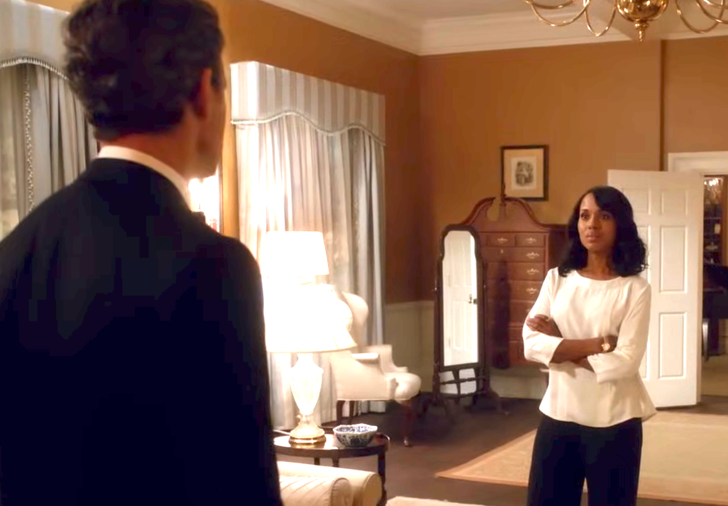 A woman stands with arms crossed facing a man in a formal suit in a well-furnished room, suggesting a tense conversation