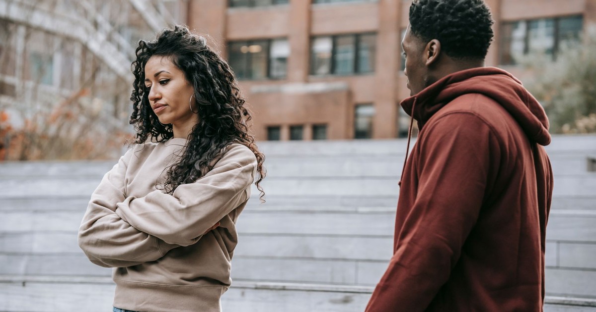 Offended ethnic girlfriend with curly hair standing with crossed arms standing near African American boyfriend while having conflict on street in park