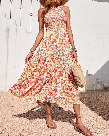 Woman in floral sundress holding straw bag, wearing a sun hat and sandals, walking on pebbled path. Perfect summer outfit inspiration