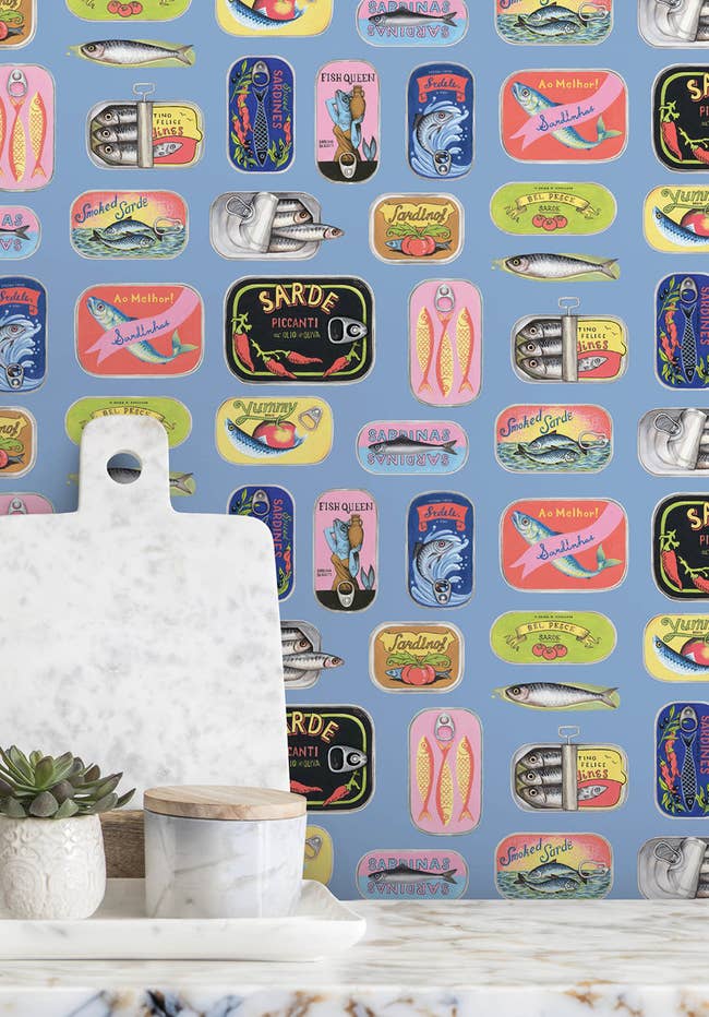 Decorative wallpaper featuring various vintage sardine can labels with a marble cutting board and potted plant in the foreground
