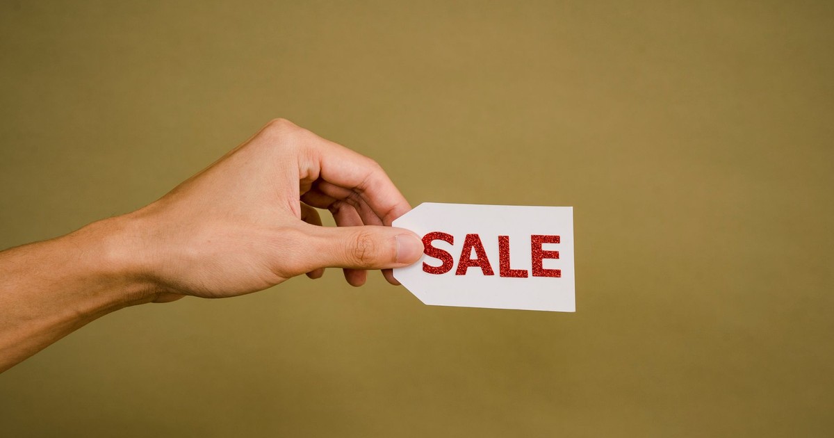 Crop anonymous person showing small white paper tag with inscription Sale against brown wall