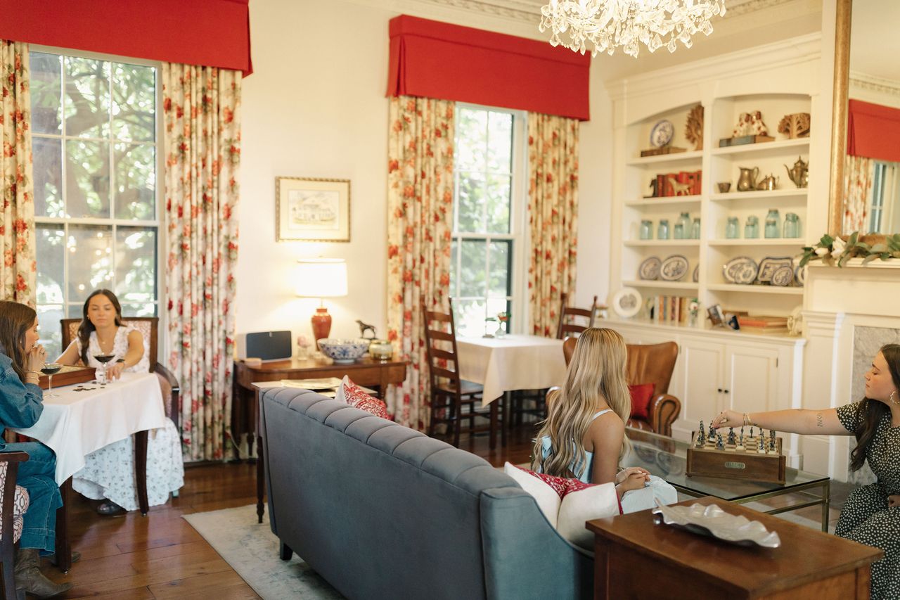 A group of people in a cozy living room with red curtains and decorative shelves sitting and conversing on a sofa and at tables