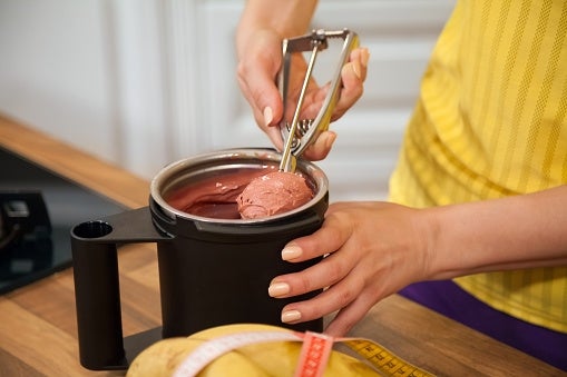 Person scooping frozen dessert from a container with a scoop. Bananas and a tape measure are on the counter nearby