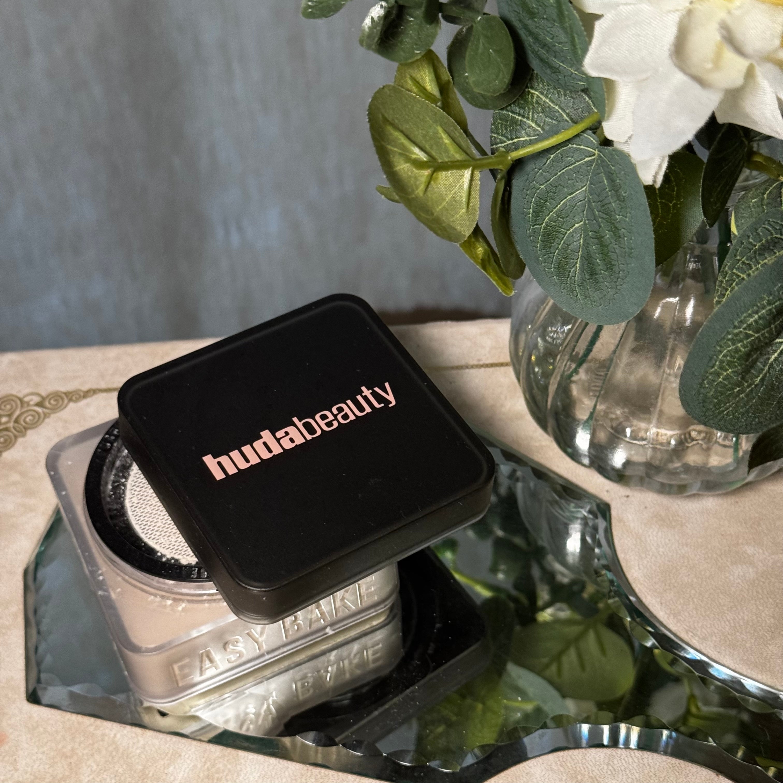 Makeup powders on a reflective surface with a decorative vase of flowers nearby