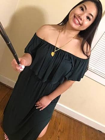 A smiling woman in an off-the-shoulder dress takes a selfie with a phone on a stick. She wears a gold necklace